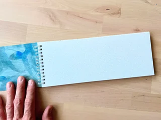 A long blue sketchbook opened flat with a hand on its painted cover and a blank white page alongside