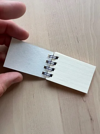 A hand holds open a miniature sketchbook, showing a white page and a ruled page joined by black wire binding