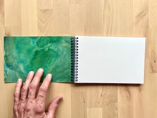 A hand holds open a handmade sketchbook with a green painted cover on the left and blank white pages on the right