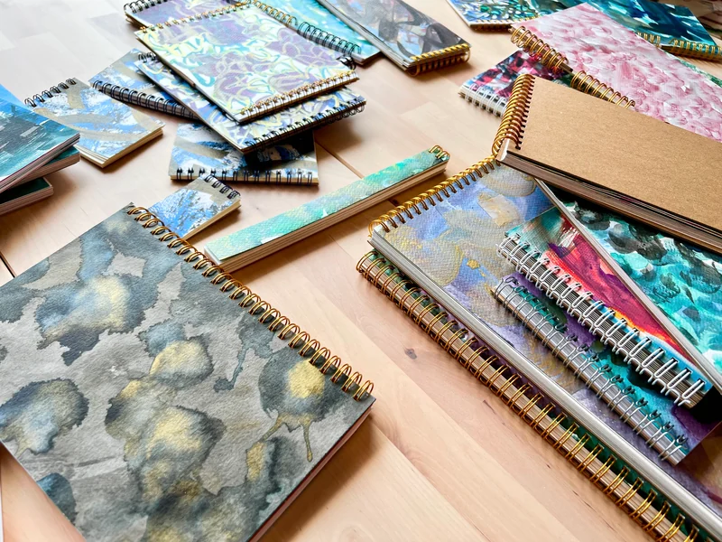 Handmade sketchbooks with painted covers, different bindings and varied formats arranged on a wooden table